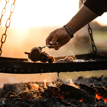 Eine Person grillt Fleisch über einem offenen Feuer bei Sonnenuntergang.