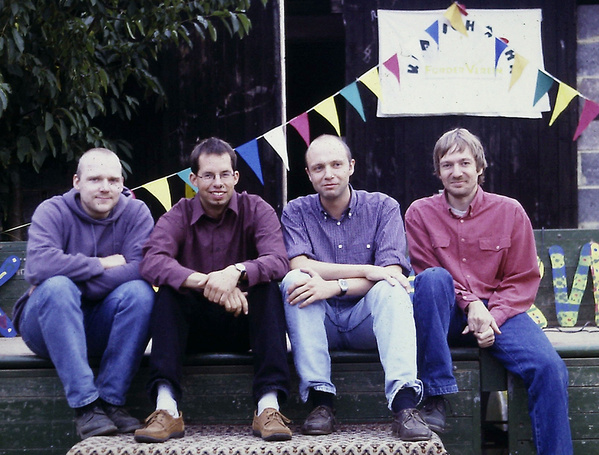 Vier Männer sitzen auf einer Bank, dahinter bunte Wimpel und ein Schild "Klatschmohn"