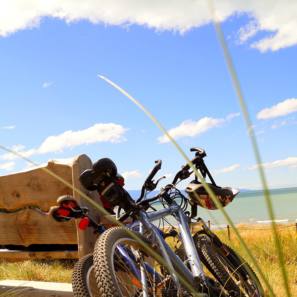 Zwei Fahrräder stehen neben einer Holzbank mit Blick auf den Strand und das Meer unter blauem Himmel.