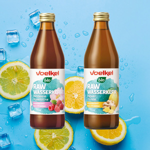  Zwei Flaschen Voelkel Bio RAW Wasserkefir vor spritzendem Wasser. Sorten: Himbeere Rosenblüte und Ingwer Zitrone.