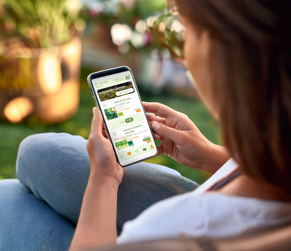 Eine Person hält ein Smartphone mit einem Gemüselieferdienst-App-Display in der Hand. Text: „hofeigenes Gemüse“.