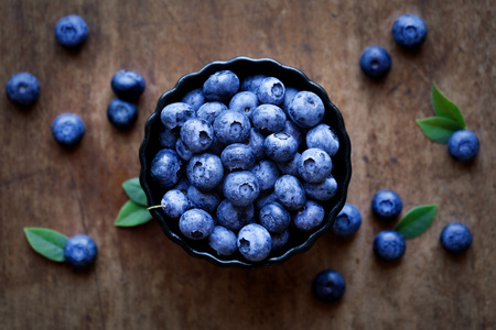 Schale voller Heidelbeeren auf einem Holztisch, umgeben von einigen losen Beeren und Blättern.