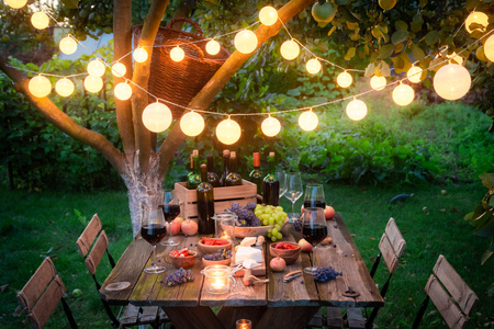 Stimmungsvoll dekorierter Gartentisch mit Wein, Käse und Lichterketten unter einem Baum.