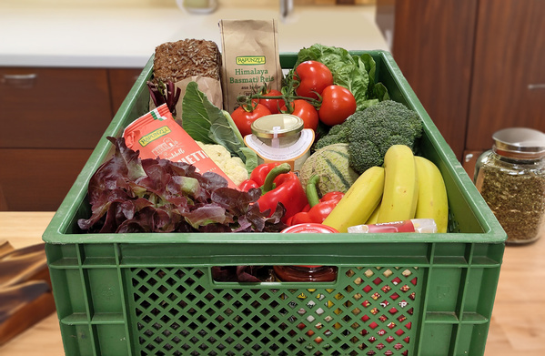 KI generiert: Eine grüne Kiste voller Obst, Gemüse und anderer Lebensmittel. Text: "RAPUNZEL, Himalaya Basmati Reis".