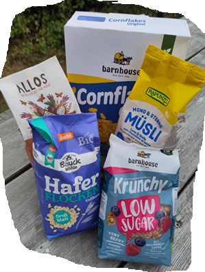 KI generiert: Verschiedene Müsliprodukte auf einem Holztisch. Enthaltene Texte: "Cornflakes", "Haferflocken", "Krunchy".