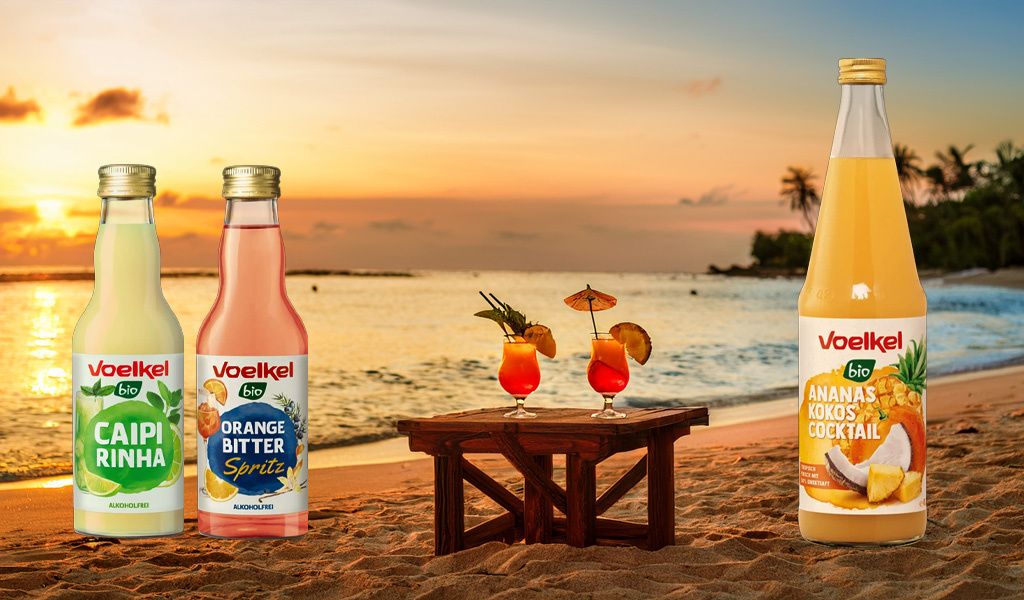  Flaschen mit Getränken vor einem Strand bei Sonnenuntergang. Text: "Voelkel bio CAIPIRINHA, ORANGE BITTER Spritz".
