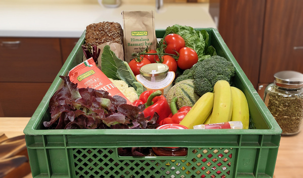 KI generiert: Eine grüne Kiste voller frischem Gemüse, Obst und verpackten Lebensmitteln. Text auf Verpackung: "Rapunzel".