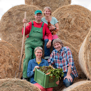 ine Gruppe von vier Personen posiert vor Heuballen. Eine hält ein Huhn, eine andere einen Gemüsekorb.