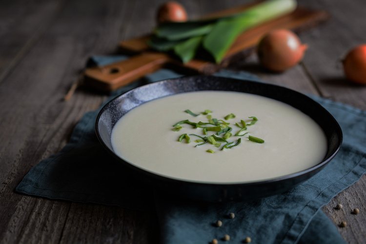 KI generiert: Ein Teller Suppe mit Kräutern, im Hintergrund Lauch und Zwiebeln auf einem Schneidebrett.