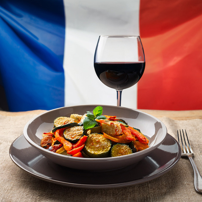 Ein Teller mit Ratatouille und ein Glas Rotwein vor einer französischen Flagge.